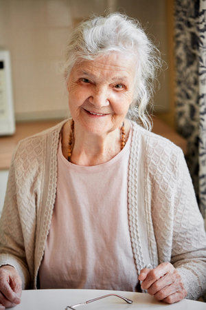 Close up portrait of elderly woman with wrinlke face and grey hair, smiling and sitting at the table...