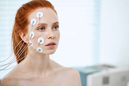 Portrait of a young red-haired woman with pony tail wearing electrodes on her face during cosmetic treatment in beauty salonの写真素材