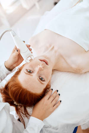 Hardware cosmetology. Close up picture of attractive woman with ginger hair and fresh freckled skin receiving rf lifting procedure in a beauty centre. Skin rejuvenation concept.の写真素材
