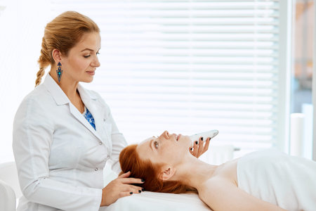 Skilled professional mature female beautician in white uniform performing photo rejuvenation...