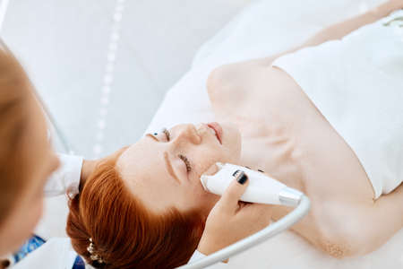 Skilled professional mature female beautician in white uniform performing photo rejuvenation cosmetology procedure for a red haired caucasian woman lying on the coach in beauty centre.の写真素材