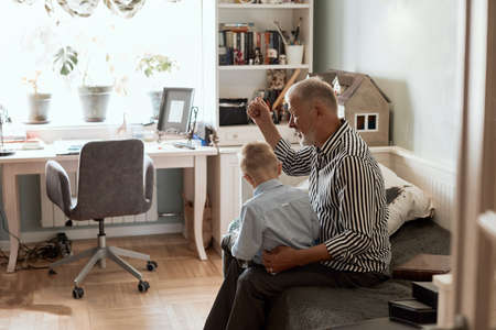 Grandson is teaching his grandfather to use digital tablet while sitting at couch in living roomの写真素材