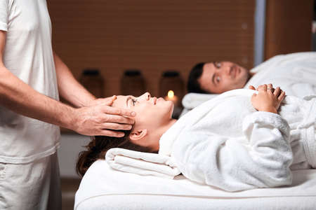 Pretty caucasian brunette girl, dressed in white bathrobe lying on table with closed eyes while having massage in spa salon. Health Care, Body Spa Concept.の写真素材