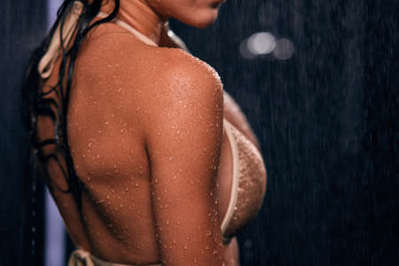 Close up photo of beautiful athletic brunette female model breast, shoulder and back with wet oiled body wearing bronze transparent sexy swimsuit posing sideways under falling water dropsの写真素材