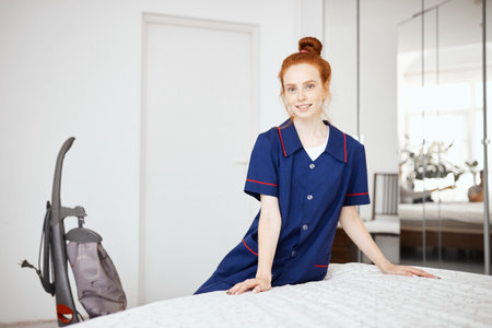 Attractive smiling ginger haired caucasian housemaid in blue working clothes looking at camera...