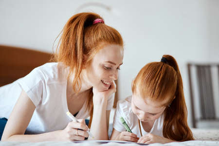 family, children and happy people concept - Close-up view of beautiful caucasian red-haired mother and daughter drawing a card for Daddy s b-day while lying on white bed, creative holydays with kids.の写真素材