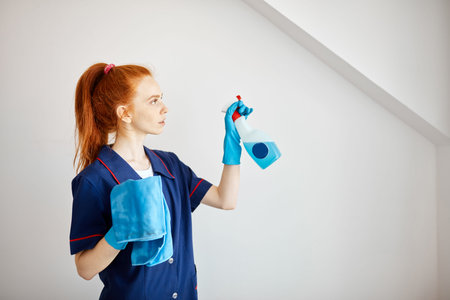 Beautiful red-haired housewife wearing household rubber gloves and blue dressing gown looking at...