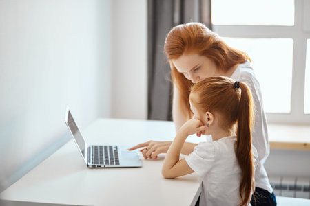 Loving caucasian red-haired mummy and her, sweetheart girl sitting at table and looking at the...