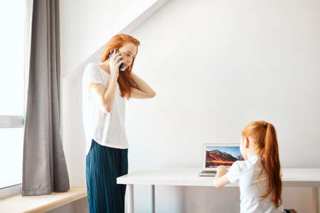 Happy redheaded mother and daughter spending time together , The little girl doing homework using laptop computer at home while her mother talking on phone at white and well-lit living roomの写真素材