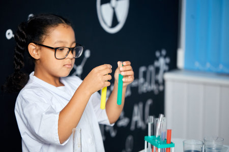 Cute female Vietnamese pupil in protective glasses and white uniform, doing biochemistry research in...