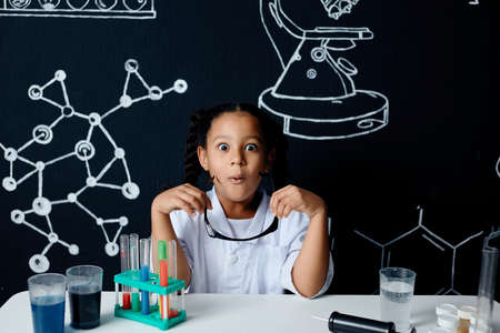 Surprised, shoked preteen girl in safety goggles and lab coat, open-mouthed and bug eyed, leaning closely to the table and watching chemical reaction going on in the bowlの写真素材