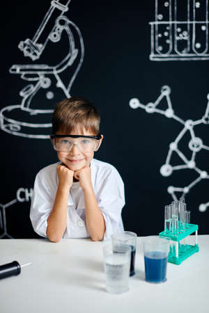 Little boy scientist in lab coat, wearing safety eyeglasses, looking at camera isolated on blackboard background with drawings and cketches.の写真素材