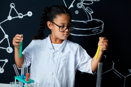 Mixed race asian, african girl in protective glasses wearing white lab holding test tubes and flasks...