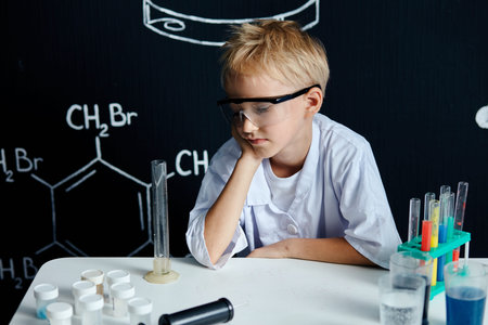 Little male scientist in white coat and protective eyeglasses smiling at camera after finishing his...