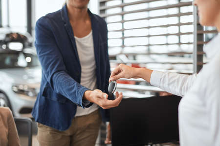 New car owners. Happy young couple receiving car keys from a salesman at the car dealership, close up. Focus on key hand giving.の写真素材