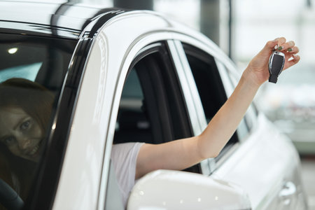 Close up of female hand holding car key while woman sits at the driver place in a car and smiling at...