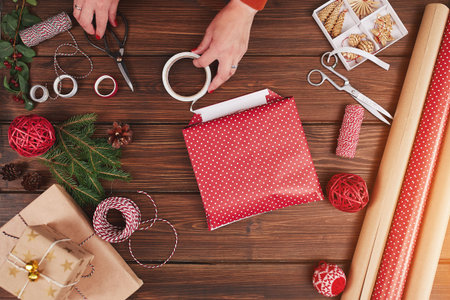 Overhead shot of female designer hands packing Christmas presents in wrapping papers on the wooden table with decorating items and Christmas baublesの写真素材