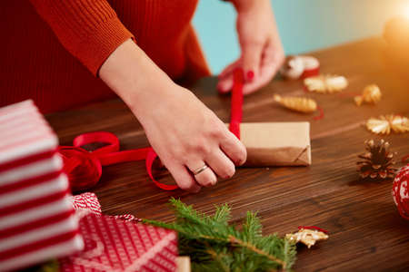 Woman s hands wrapping Christmas presents in colored and paper decorated with red ribbon, fir branches and pine cones on a rustic wooden board. Top viewの写真素材