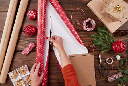 Woman in a red sweater wrapping gift box with equipment and decorating items on wood table, preparing for celebrating Christmas holidays - top view.の写真素材
