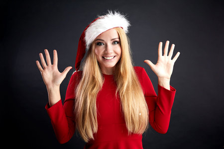 Excited young blonde girl with long hair wearing Santa hat and red elegant dress gesturing with two...