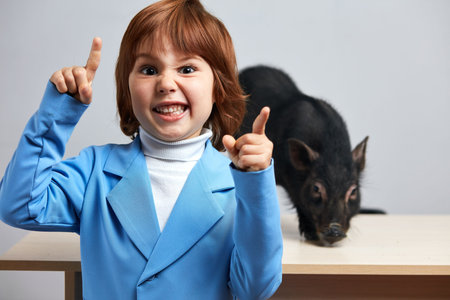 Indoor portrait of little red-haired boy in blue jacket with his exotic pet - little black pig,...