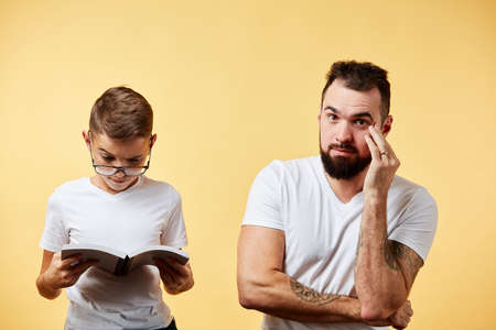 father incredulously looking at cute son reading book at yellow backgroundの写真素材