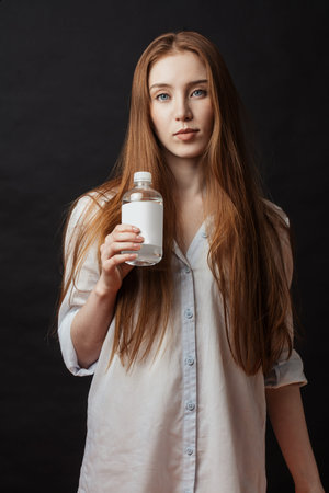 Studio shot of tender young female model with long loose hair holding bottle of water, isolated on...