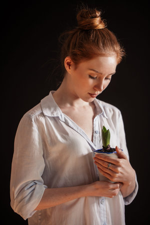 Young attractive woman with ginger hair bun in white shirt holding potted plant isolated ober black...