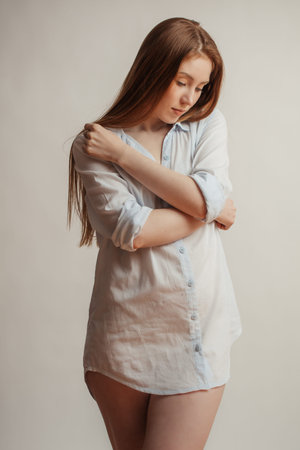 Young female model with long loose red hair embracing herself with hands as if she is shy,...