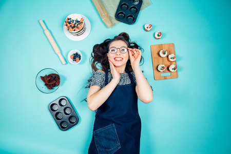 Top view of happy housewife with perfect smile lying on a blue background with cake and bakery gadgets aroundの写真素材