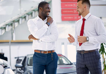 confident caucasian man salesperson selling cars at car dealership, he is presenting special offers to african american clients, have conversationの写真素材