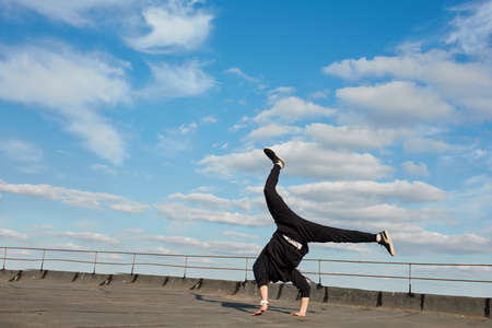 Asian hip-hop man dancer dressed in black sports pants, sweatshirt jumping in air, making a backdancing stunts outdoor on the roof over blue sky backgroundの写真素材