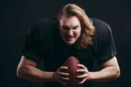 Emotional portrait of strong caucasian American football player with aggresive look and unprotected bare head in black team uniform prepares to throw the ballの写真素材