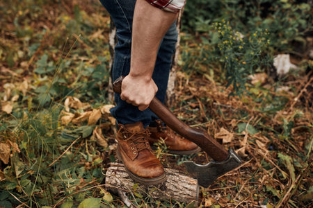 Man in jeans and checkered shirt holding old axe on stump with leg