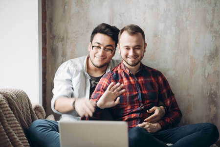 Romantic partners in love and business. Caucasian amused gay couple looking at laptop screen while resting in living room with loft interior. Dominant man wearing spectacles and and using laptopの写真素材