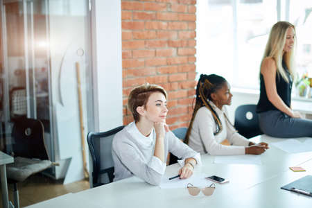 fair-haired beautiful stylish girl sitting at the table with pensive look and listening to her colleagues in the modern office with loft design . close up side view shotの写真素材