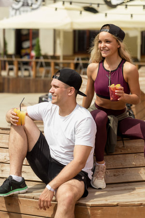 Sportive man and attractive blond woman in tracksuit sitting in outdoor cafe in summer morning with...