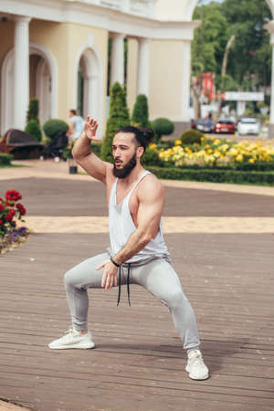 Caucasian athetic man performing squats exercise training in summer park with building on backgroundの写真素材