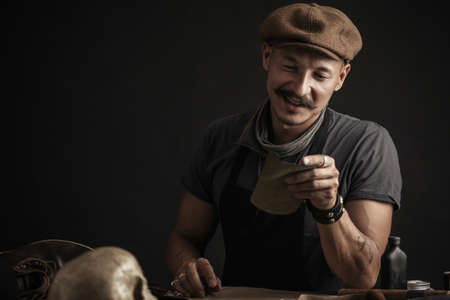 Whiskered handsome shoemaker wearing stylish cap working with leather, making measurments in patterns at table in workshop studio.の写真素材