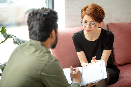 young woman is sitting on couch and talking to male indian psychologist while doctor making notes