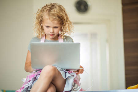 Little business lady. Cute caucasian girl sitting at the table and using laptop, listening sound in headphones not to disturb her sleeping parentsの写真素材