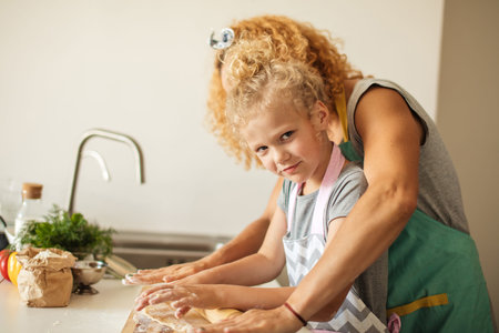 Little frowning female Chef with the help of Mom preparing dough and ingredients for pizza, looking...