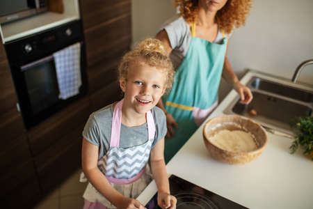 High angle view of caucasian pretty young woman and her little cute daughter are cooking at the kitchen, girl is at focus, looking at camera.の写真素材
