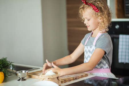 Cute little girl with red headband on frizzy blonde hair wearing apron, making the dough for vegetable pie while preparing it for baking.の写真素材