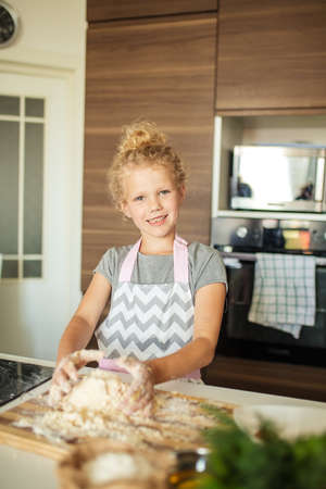 Cute little caucasian girl with blonde curly hair tied in ponytail in apron looking at camera and baking cookies at home kitchenの写真素材