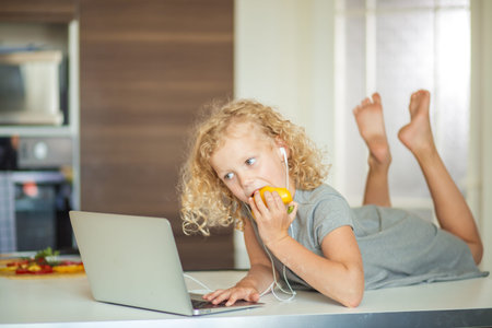 Little curly girl in home wear eating paprica and playing computer game using laptop and earpods in...