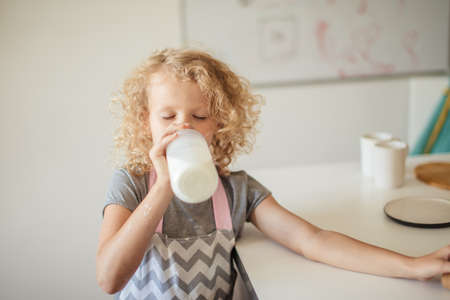 Cute little curly blonde small girl drinling milk on white background while helping mother in the kitchen. Health and Child Beauty conceptの写真素材