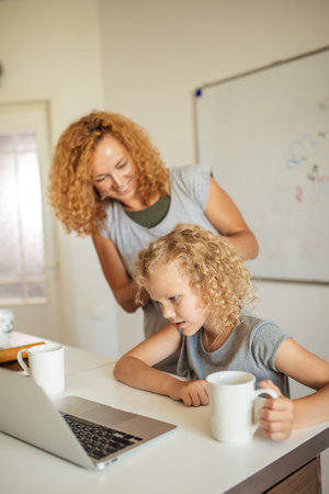 Happy young blonde mother ties up gorgeous frizzy hair of her daughter while the adorable female kid...