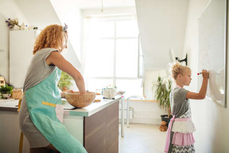 Schoolgirl in home wear and apron writing something on white board on the wall with a marker and her mother looking at her. Learning and school concept.の写真素材