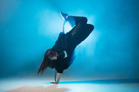 Young hip-hop female dancer performing solo on the stage in blue neon lights with flash light in the...